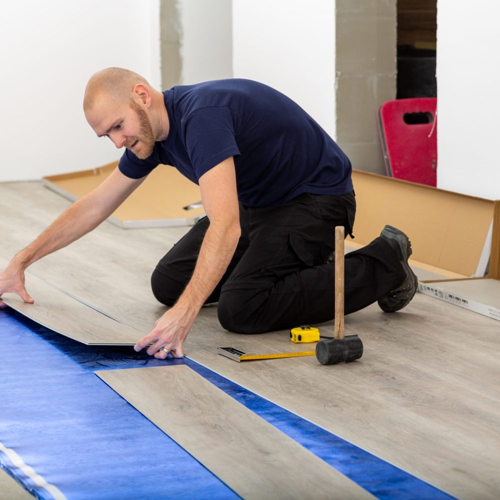 Worker joining click lock vinyl floor plank covering at home renovation. Man laying PVC-floor. Craftsman installing new laminated wooden floor on blue acoustic thermal flooring underlayment.